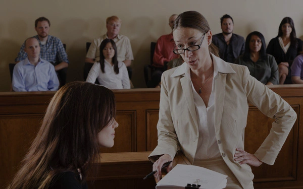 A woman in a beige blazer is speaking while holding papers in a courtroom; another woman sits opposite her. Several people are seated in the background, observing.