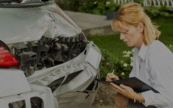 A woman examines and takes notes on a damaged car with a broken rear bumper.