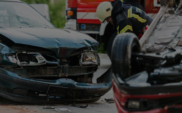 A firefighter inspects two damaged cars, one upright and one overturned, with a fire truck visible in the background.