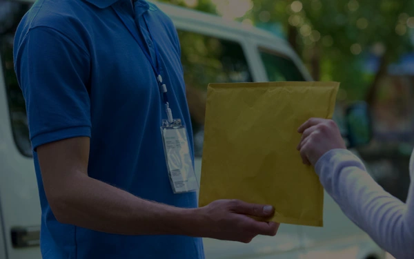 A person in a blue polo shirt receives a yellow envelope from another person. A van is seen in the background.