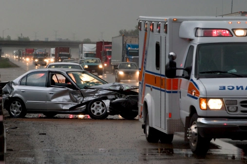 A damaged car is in the road after an accident, with an ambulance nearby. Traffic is backed up in the opposite direction. It's overcast with wet road conditions.