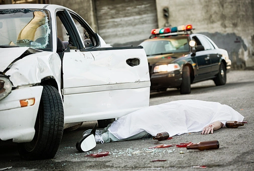 Car crash scene with a damaged white vehicle, a person covered by a white sheet on the ground, broken glass, and beer bottles. A police car with flashing lights is in the background.