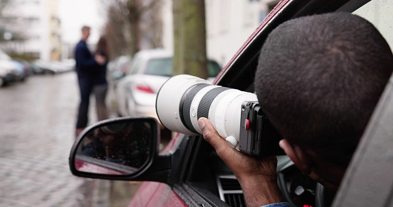 Person sitting in a car holds a large camera lens, photographing a couple standing on a street.