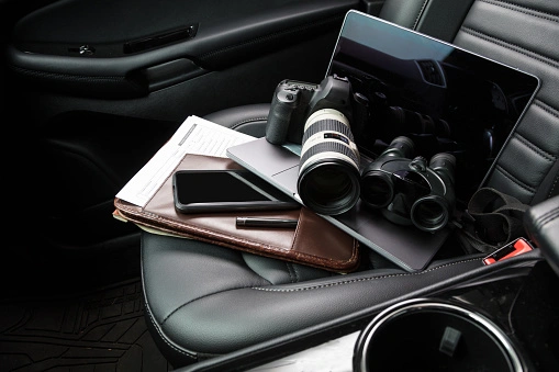 A car seat holding a camera, laptop, binoculars, smartphone, papers, and a pen.