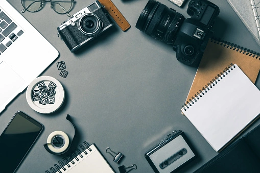 A flat lay of photography and office equipment, including cameras, a laptop, notebooks, and a smartphone on a gray desk.