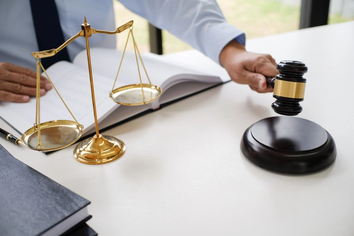 A judge's hand holds a gavel next to a book and a set of scales on a desk.