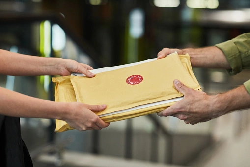 Two people exchange a stack of sealed yellow envelopes indoors.