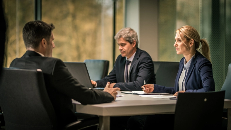 Three professionals in a meeting room, two men on one side of a table and a woman on the other side, all wearing business attire and engaging in discussion.